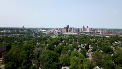 Wide-aerial-shot-tilting-down-and-showing-the-city-of-Saint-Paul,-Minnesota-on-the-horizon-with-residential-houses-and-trees-in-front-and-a-clear,-blue-sky-above