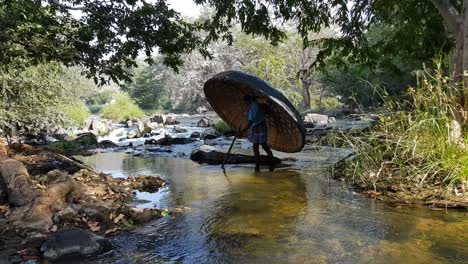 A-man-crossing-a-river-stream-of-the-Cauvery-river-carrying-a-coracle-and-a-paddle-in-Hogenakkal,-Tamilnadu,-India