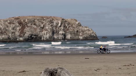 Two-ladies-ride-there-bikes-on-a-beach-in-Bandon,-Oregon