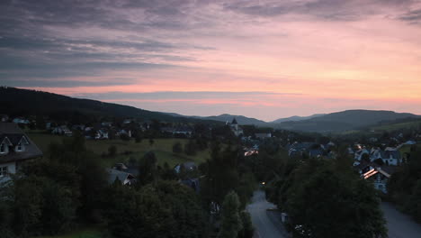 Wide-panoramic-pan-over-the-mountainous-spa-village-of-Grafschaft-in-the-winter-sports-region-of-Sauerland,-Germany,-during-sunset-with-a-colourful-purple-and-orange-lit-cloudy-sky