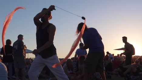 Close-Up-of-a-Man-Performing-with-Poi-Ribbons-at-a-Music-Festival-with-the-Sun-Setting-in-the-Background