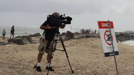 Ein-Nachrichtenkameramann-Neben-Einem-Warnschild,-Das-Videoaufnahmen-Von-Schäden-An-Einem-Strand-Durch-Einen-Sich-Entwickelnden-Zyklon-Macht