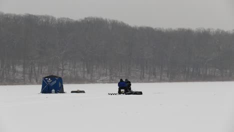 Two-men-sitting-on-chairs-in-the-middle-of-a-frozen-lake-during-blizzard-conditions-are-ice-fishing