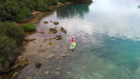 Aerial-view-of-woman-stand-up-paddle-by-shoreline-in-clear-turquoise-water-by-tropical-island