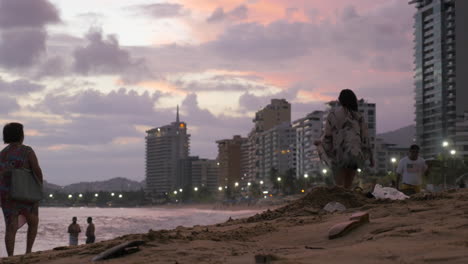 People-walking-along-beach-in-Acapulco,-Mexico-at-sunset-with-city-background