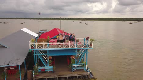 Eine-Gruppe-Von-Freunden-Winkt-In-Die-Kamera,-Während-Sie-Auf-Einem-Hausboot-Auf-Dem-Tonle-Sap-See-In-Kambodscha-Steht-Und-Die-Kamera-Rückwärts-Fliegt