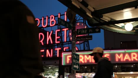 Street-and-neon-sign---people-outside-the-Public-Market-Center-and-Pike-Place-Fish-Company---Dark-moody-sky-in-the-background,-birds-and-street-lights