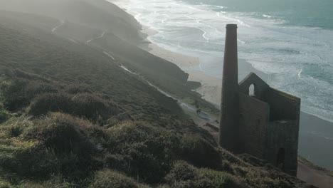 Cornish-tin-mine-pump-house-on-the-north-Cornwall-coast-with-people-walking-the-coastal-paths-and-the-beach