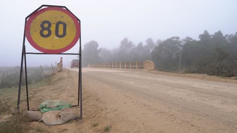 Roter-Lieferwagen-Fährt-Im-Nebel-Auf-Einer-Unbefestigten-Straße-Auf-Dem-Land-An-Einem-Geschwindigkeitsschild-Mit-80-Km/h-Vorbei-über-Eine-Brücke