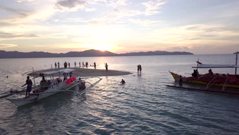 Vista-Aérea-De-ángulo-Bajo-De-Un-Turista-Disfrutando-De-La-Puesta-De-Sol-En-Una-Pequeña-Isla-En-Palawan,-Filipinas-Con-Dos-Barcos-Locales