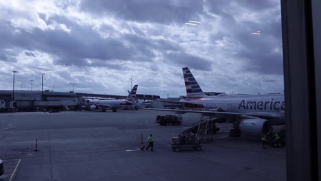 Airplanes-at-airport-gate,-workers-and-small-truck-and-cargo-drive-by-on-cloudy-grey-day,-static