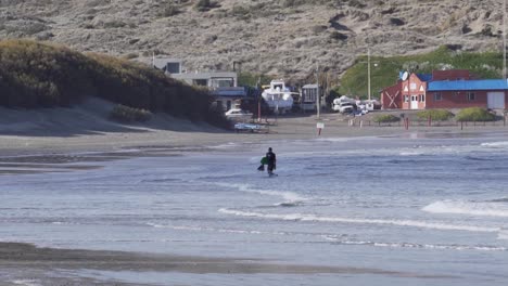 Man-Holding-A-Body-Board-Walking-Out-Of-The-Rough-Sea-On-A-Sunny-Summer-Day---wide-slowmo-shot