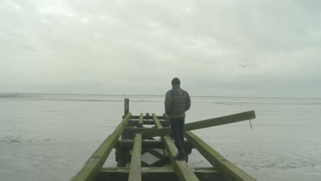 Guy-in-green-rain-jacket-looks-into-the-distance-with-birds-flying-past-the-clouds-over-the-north-sea-near-Husum,-Germany,-Slow-Motion