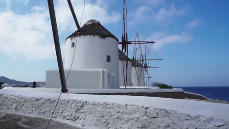 Panorámica-De-Los-Famosos-Molinos-De-Viento-De-Mykonos-Mientras-Un-Pájaro-Intenta-Aterrizar-En-El-Techo,-Con-Turistas-Caminando-Alrededor-De-Ellos-Hasta-El-Mar-Egeo.