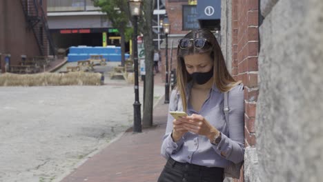 A-millennial-girl-looks-at-phone-texting-friends-wearing-a-face-mask-during-a-pandemic,-leaning-against-the-building