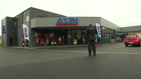 Toma-De-La-Entrada-Del-Supermercado-Mientras-Pasan-Automóviles-Y-Clientes,-Con-Hombres-De-Seguridad-Controlando-El-Tráfico