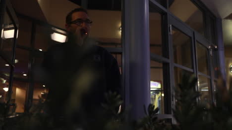 Man-on-a-cold-night-smoking-while-he-is-standing-outside-of-a-pub,-club-or-hotel-while-he-is-looking-into-the-distance