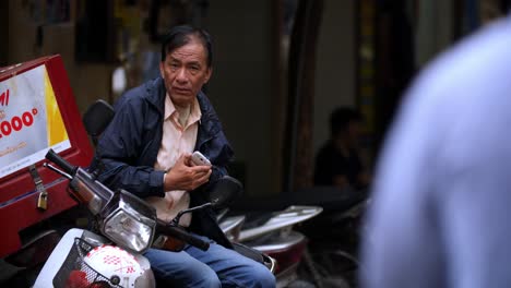 Man-pulls-out-cellphone-from-his-pocket-and-turns-it-on-while-sitting-sideways-on-a-motorbike-parked-on-the-street-in-daytime,-Handheld-slow-motion-shot