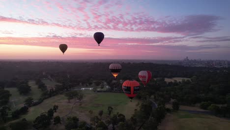 Drohnenaufnahmen-Von-Heißluftballons,-Die-Bei-Sonnenaufgang-über-Grünen-Parks-Mit-Der-Stadt-Melbourne-Im-Hintergrund-Abheben