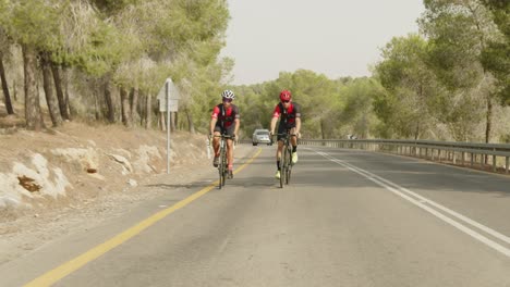 slow-motion-wide-shot-of-two-road-cycelist-professionals-riding-near-a-forest-while-a-car-behinds-them