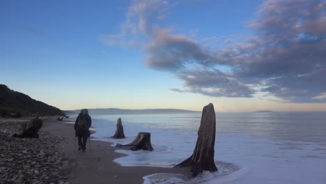 Hiker-with-backpack-and-poles-walks-on-remote-ocean-beach-at-dawn