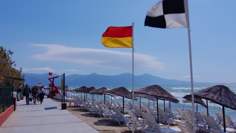 Junger-Schwarzer-Mann-Schlendert-Mit-Helmen-In-Zeitlupe-Entlang-Einer-Strandpromenade-An-Der-Türkischen-Küste,-Während-Die-Fahnen-Im-Wind-Flattern