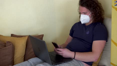 Young-man-with-a-mask-sitting-on-his-bed-sends-a-message-on-his-cell-phone-then-continues-working-on-his-laptop