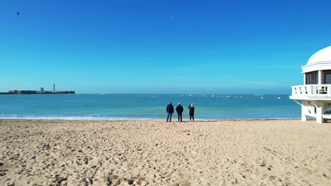 Luftaufnahme:-Drei-Menschen-In-Schwarzer-Kleidung-Stehen-Am-Strand,-Während-Vögel-Um-Den-Strand-Herumfliegen