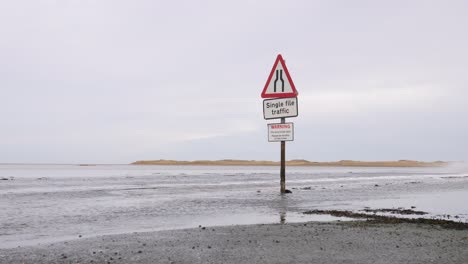 Red-car-driving-along-flooded-causeway-past-warning-sign