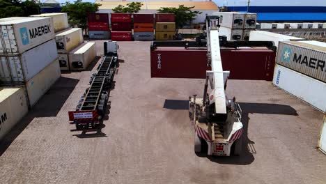 Heavy-machinery-crane-transporting-red-shipping-container,-rear-aerial
