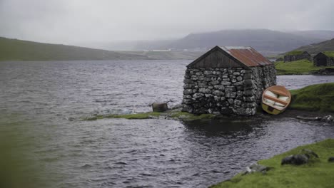 Wide-shot-of-old-stony-abandoned-building-at-shore-of-natural-lake-during-cloudy-day-on-Faroe-Islands