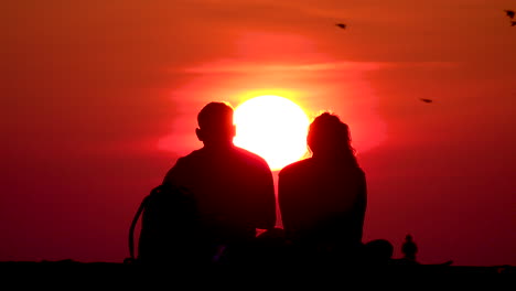 Ein-Paar-Sitzt-Am-North-Avenue-Beach-In-Chicago-Vor-Einer-Leuchtend-Gelben-Sonne-Mit-Orangefarbenem-Himmel,-Während-Ein-Vogelschwarm-Vorbeifliegt