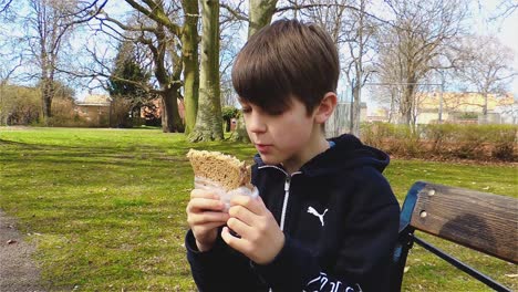 Niño-Hambriento-Comiendo-Un-Sándwich-En-Un-Banco-Del-Parque-Y-Disfrutando-Del-Aire-Libre