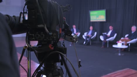 A-close-look-behind-a-cameraman:-an-operator-is-setting-up-a-camera-and-filming-a-talk-between-5-men-seated-in-modern-chairs