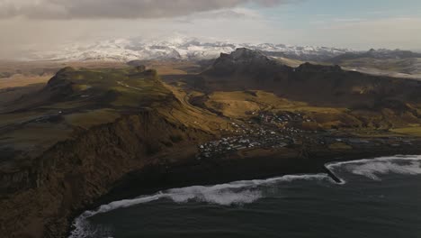 Aerial-footage-showcasing-the-town-of-Vik,-Iceland,-with-its-dramatic-coastal-cliffs-and-snow-capped-mountains-in-the-background