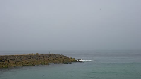 Fisherman-standing-on-breakwater,-fishing-on-the-rocky-shore-with-a-view-of-the-misty-sea