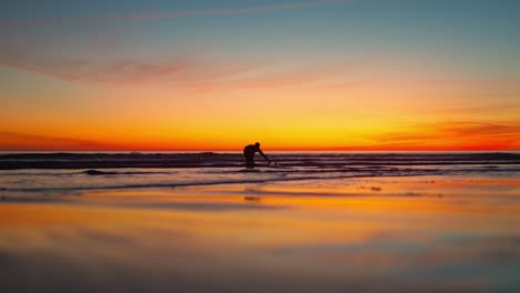 Plano-Amplio-De-Una-Surfista-Lavando-Una-Tabla-De-Surf-Al-Atardecer-Y-Caminando-De-Regreso-A-La-Playa-En-Costa-Da-Caparica,-Portugal