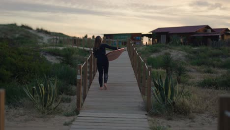Medium-Shot-of-Female-Surfer-Walking-on-Wooden-Pathway-with-Surfboard-at-Dusk-in-Costa-da-Caparica,-Portugal