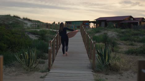 Plano-Medio-De-Una-Surfista-Caminando-Por-Un-Sendero-De-Madera-Con-Una-Tabla-De-Surf-Al-Atardecer-En-Costa-Da-Caparica,-Portugal