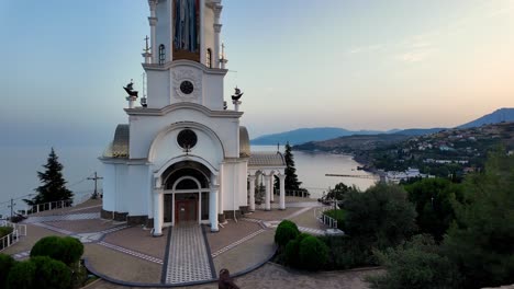 A-beautiful-white-church-with-a-bell-tower-overlooking-the-sparkling-sea-and-lush-green-hills-at-sunset-in-Crimea