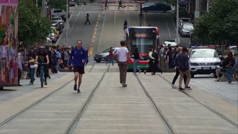 Melbourne-urban-lifestyle,-tram-running-along-Bourke-street,-passing-through-a-bustling-city-with-pedestrians-crossing,-commuters-waiting-at-the-stop-and-skater-boy-manoeuvring-through-the-street