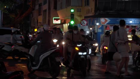 Turistas-Con-Cascos-Esperan-Por-La-Noche-En-Una-Concurrida-Intersección-De-Hanoi-Llena-De-Motocicletas-Y-Luces.