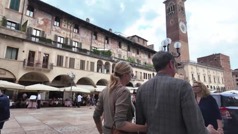 Famoso-Monumento-Piazza-Delle-Erbe-Con-Mercado,-Restaurantes,-Reloj,-Torre,-Gente,-Autos,-Pareja-Tomados-De-La-Mano,-Verona,-Italia