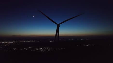 A-wind-turbine-stands-against-a-twilight-sky,-city-lights-twinkling-below
