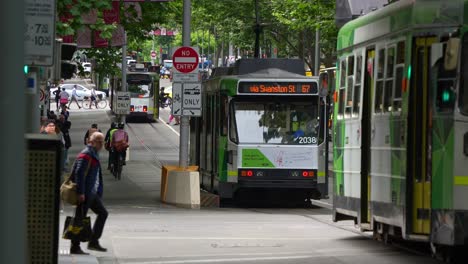 Trams-glide-along-Swanston-Street-in-Melbourne's-bustling-central-business-district,-as-pedestrians-cross-at-the-crosswalks,-showcasing-the-hustle-and-bustle-of-Australian-urban-lifestyle