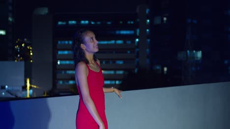 On-a-rooftop,-a-Hispanic-girl-stands-with-the-luminous-city-lights-of-Port-of-Spain,-Trinidad-in-the-Caribbean-background