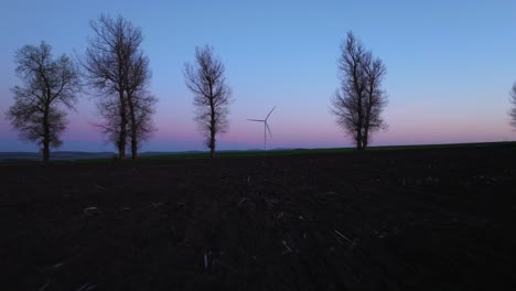 Wind-turbine-and-bare-trees-silhouetted-against-a-vibrant-dusk-sky-in-a-tranquil-rural-landscape