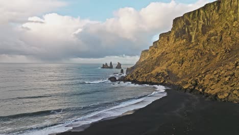 Aerial-footage-of-the-rugged-coastal-cliffs-and-iconic-sea-stacks-near-Vik,-Iceland,-with-waves-crashing-against-the-shore