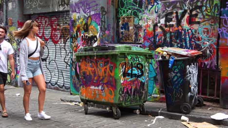 Tourists-visiting-Hosier-Lane,-a-popular-cobblestone-laneway-featuring-a-vibrant-array-of-art-murals-and-graffiti-on-the-exterior-walls-of-buildings-with-overflowing-rubbish-from-the-wheelie-bins