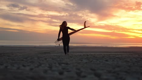 Plano-Amplio-De-Una-Surfista-Caminando-Por-La-Playa-Con-Una-Tabla-De-Surf-Al-Atardecer-En-Costa-Da-Caparica,-Portugal